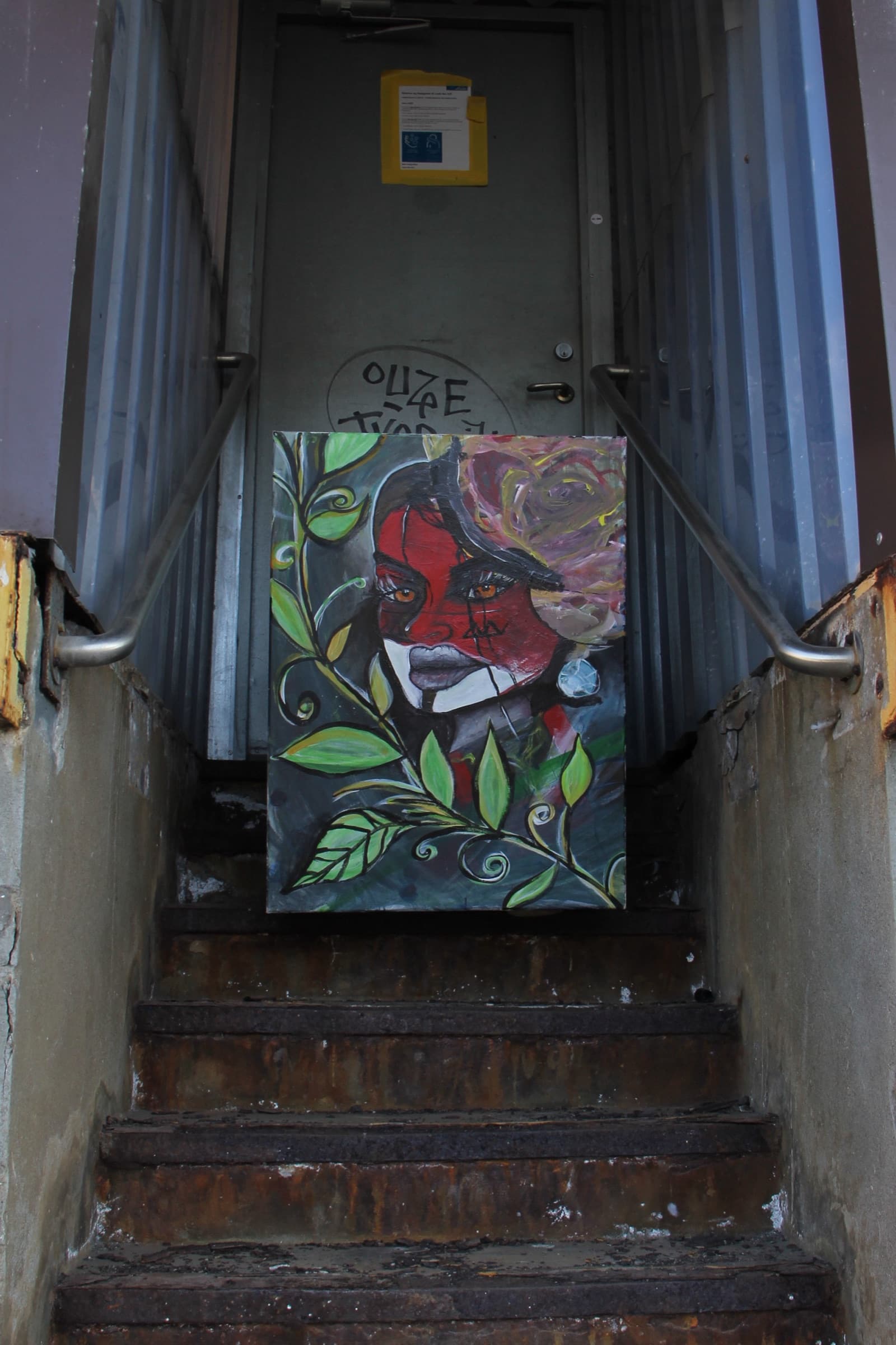 The portrait standing at the top of a narrow concrete stairwell in front of a tagged metal door