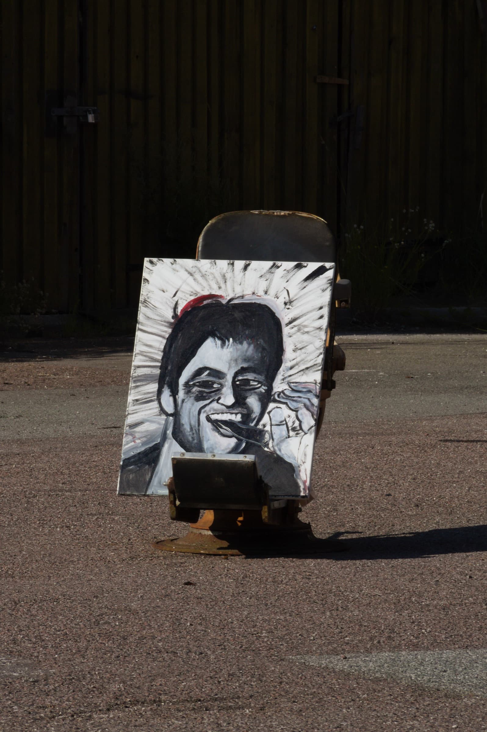 The painting propped on a rusted swivel seat in an empty lot, lit by low sun against a dark timber fence